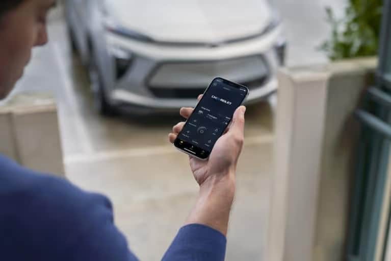 Person using a smartphone to control a smart car in a driveway surrounded by greenery, showcasing modern technology and convenience in a residential setting.