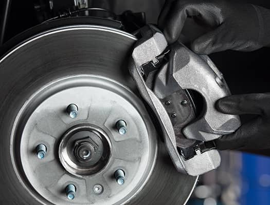 Close-up of hands wearing black gloves holding a metallic brake caliper next to a silver brake disc with lug nuts visible. The image shows part of a vehicles wheel assembly.