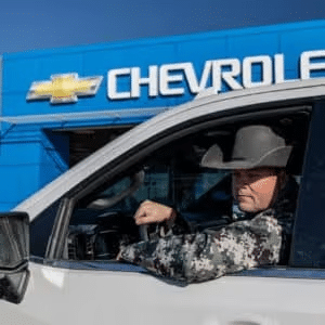 Texan cowboy wearing a hat and camo jacket sitting in the driver's seat of a white Chevrolet truck, with a blue Chevrolet dealership sign in the background.