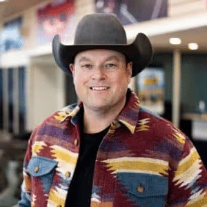 Smiling man wearing a cowboy hat and a colorful patterned jacket, standing inside a modern interior space with promotional materials in the background.