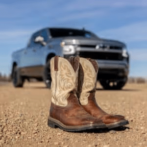 A pair of worn cowboy boots with decorative designs in the foreground, with a shiny silver pickup truck parked on a dirt road in the background under a clear blue sky.