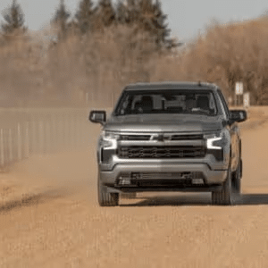 Silver Chevrolet truck driving on a dirt road, kicking up dust with trees in the background. Ideal for showcasing off-road capability and rugged design.
