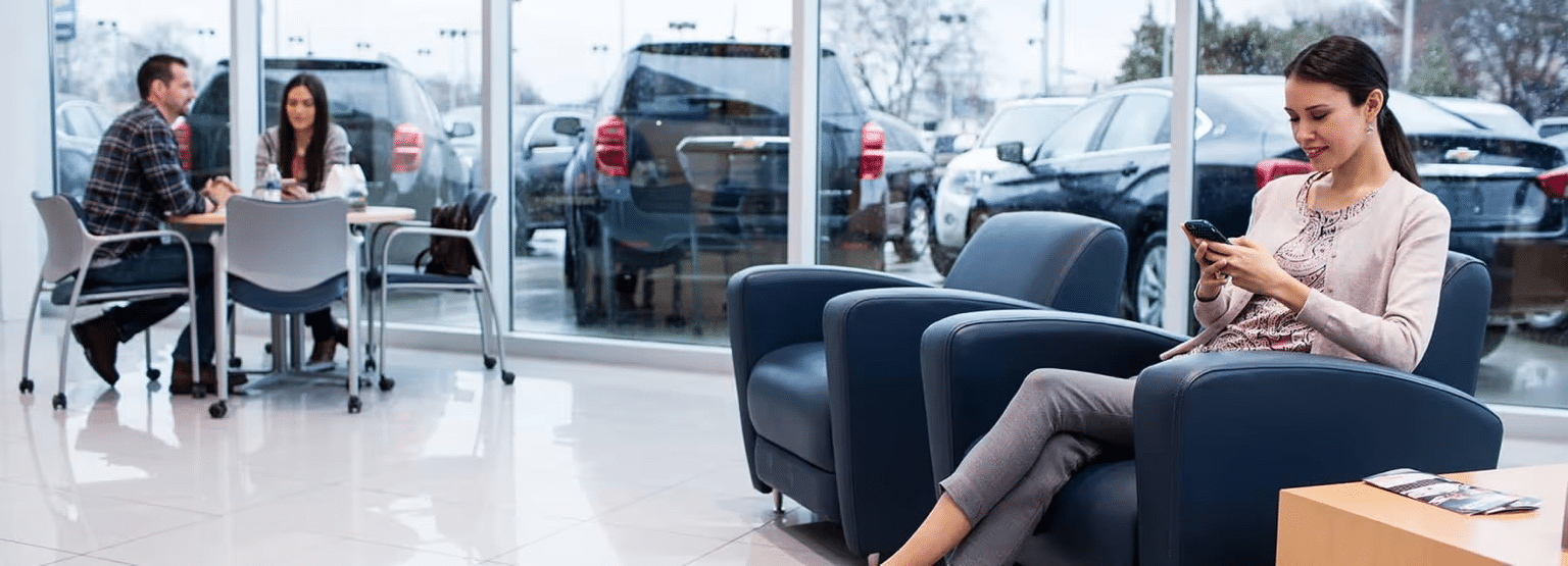 A woman casually texting on her phone while seated in a modern car dealership waiting area, with a couple engaged in conversation at a nearby table and parked vehicles visible through large glass windows.