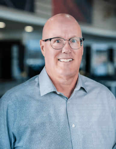 Portrait of a smiling middle-aged man with glasses, wearing a light gray button-up shirt, set against a modern office background with soft lighting.
