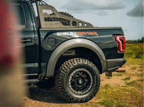 Close-up view of a black Shelby F-150 truck, showcasing its rugged tires, custom wheel design, and distinctive rear branding in an outdoor setting.