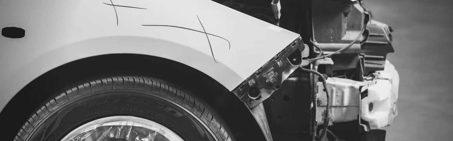Close-up of a car's damaged front fender with a visible scratch mark, highlighting the tire and wheel. The image shows a section of the car body exposed, indicating repair work in progress.