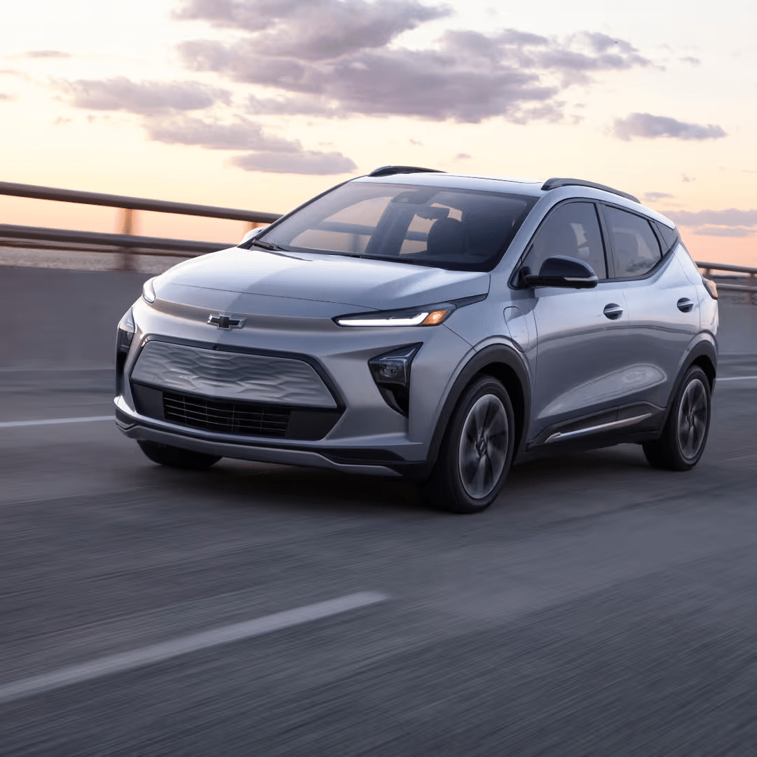 Silver Chevrolet Bolt EV driving on an open roadway during sunset, showcasing its sleek design and modern features.