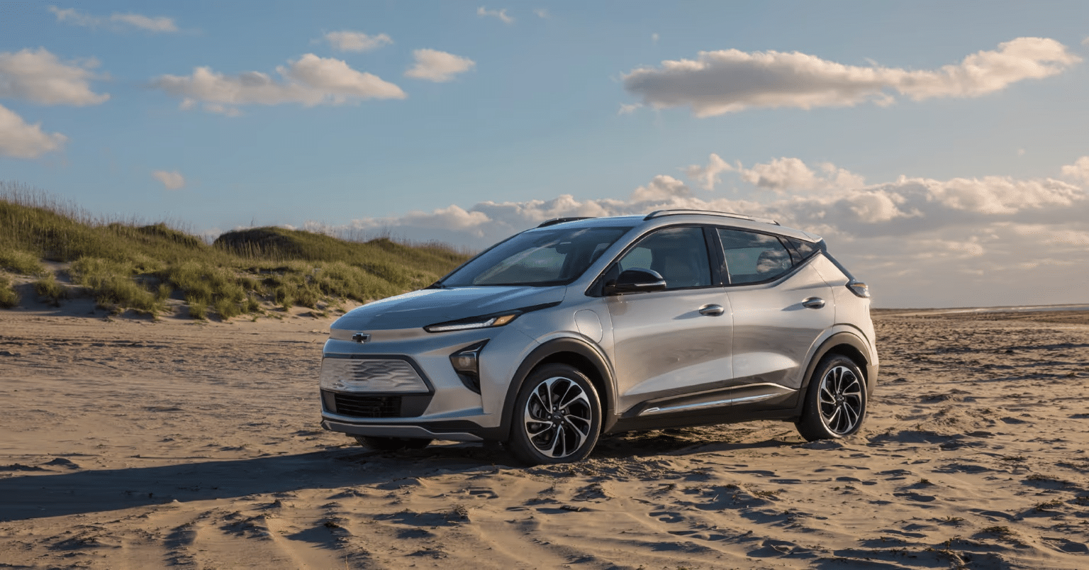 A silver Chevrolet electric vehicle parked on sandy beach terrain with grass dunes and a blue sky in the background.