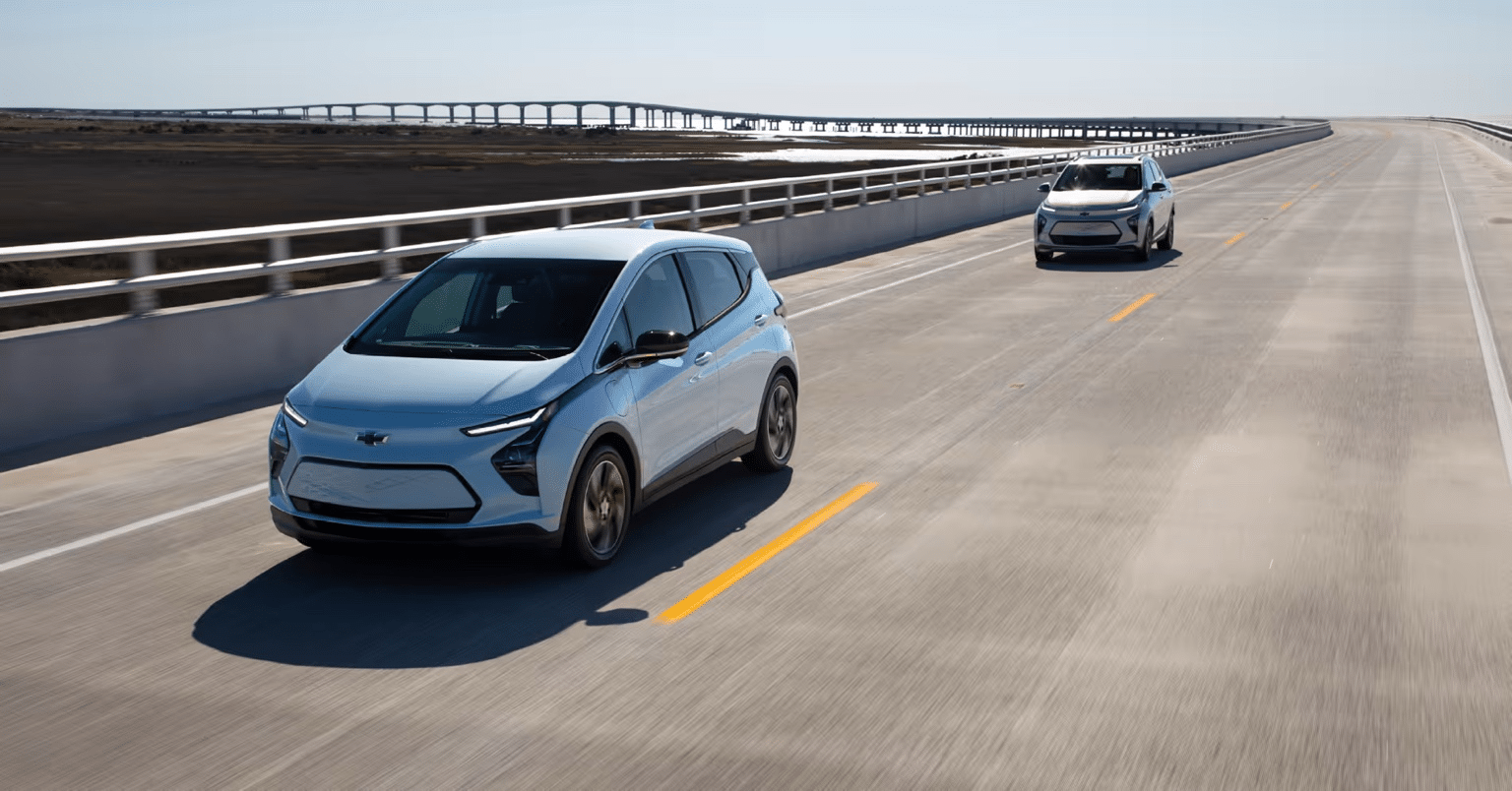 Two Chevrolet electric cars driving on a wide, modern highway with a bridge in the background, showcasing the appeal of electric vehicles and scenic routes.