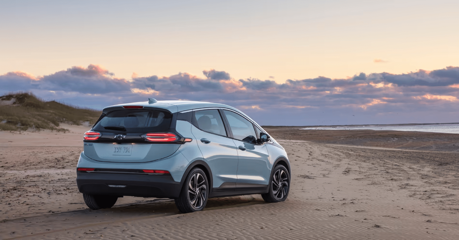 Electric Chevrolet Bolt EV parked on a sandy beach at sunset, showcasing its sleek design against a backdrop of clouds and coastline.
