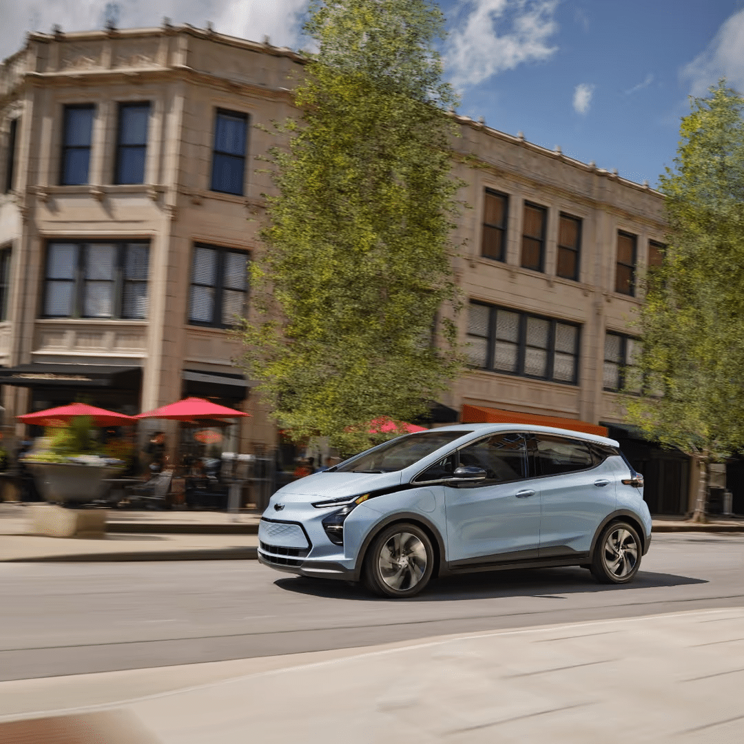 Electric blue vehicle driving through an urban streetscape with historic buildings and outdoor dining areas, showcasing modern design and eco-friendly transportation.