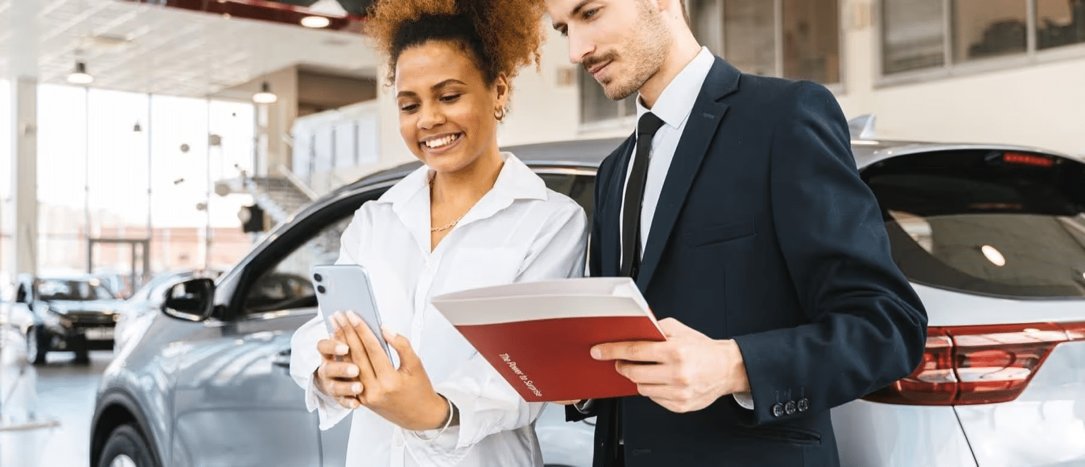 Sales representative and a customer discussing vehicle features in a modern car dealership, showcasing a friendly interaction with a smartphone and vehicle brochure.