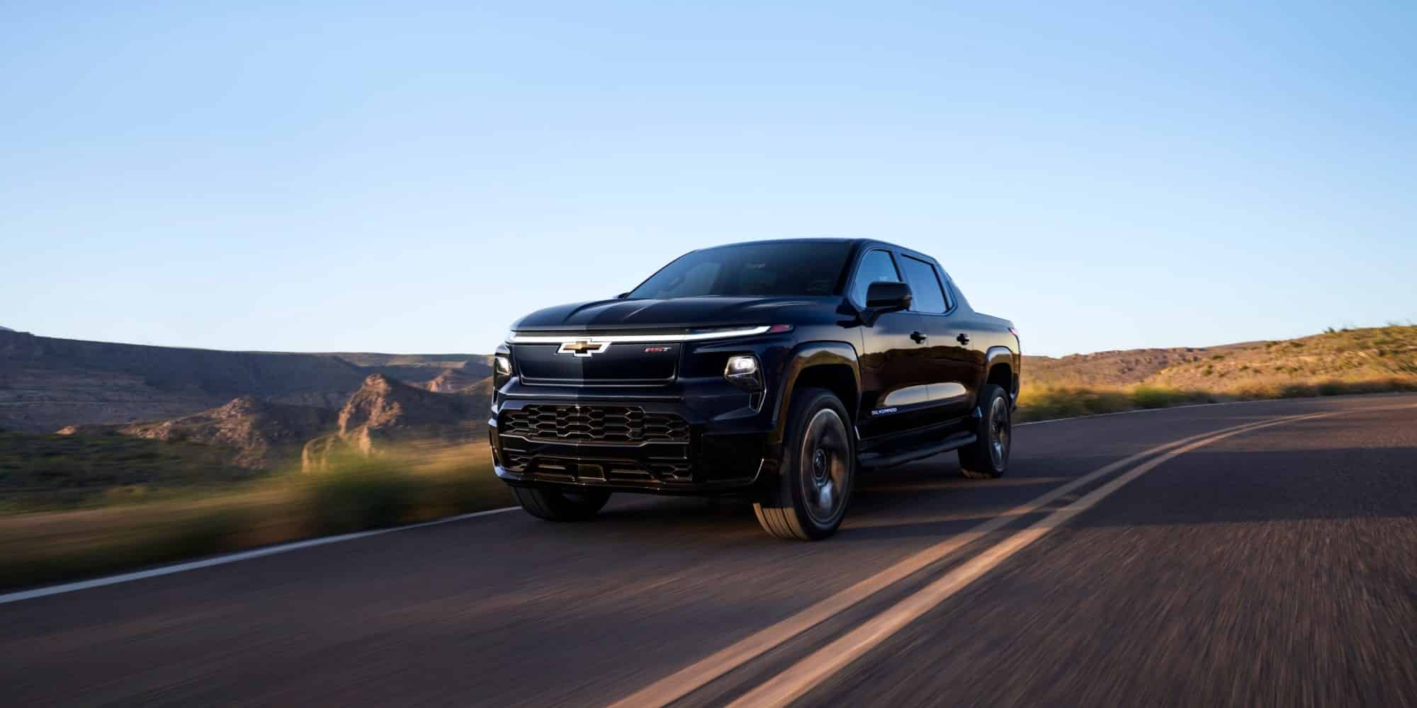Black Chevrolet Silverado EV driving on a winding road in a scenic mountainous landscape, showcasing its sleek design and electric capabilities.